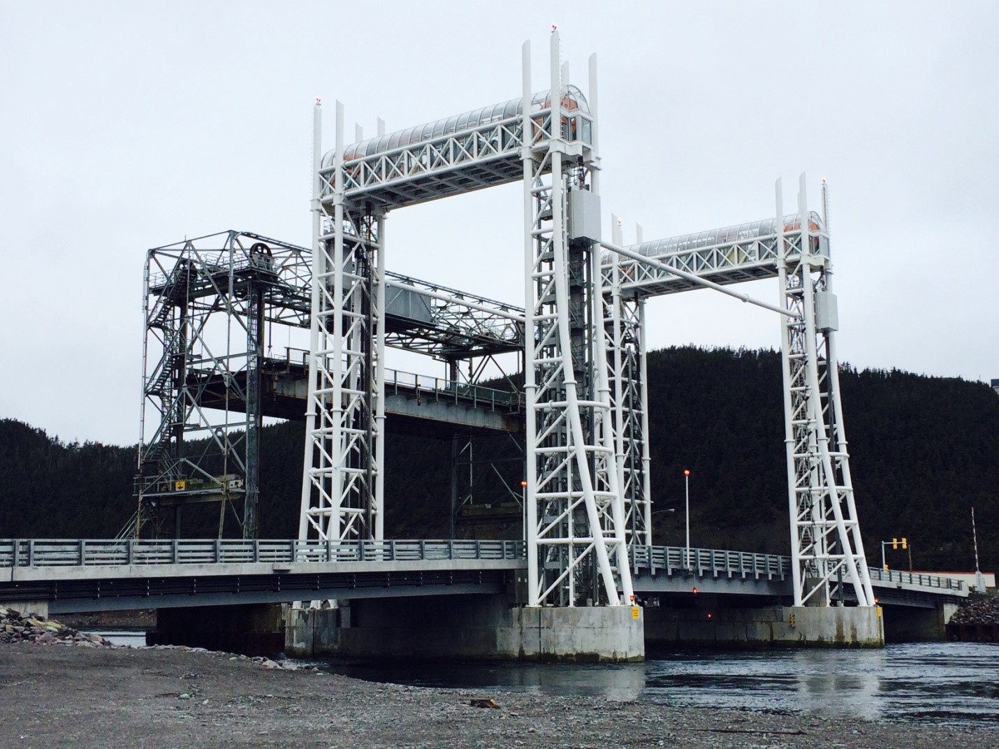 A large vertical lift bridge with white steel towers stands over a body of water, with a forested hillside in the background under a cloudy sky.