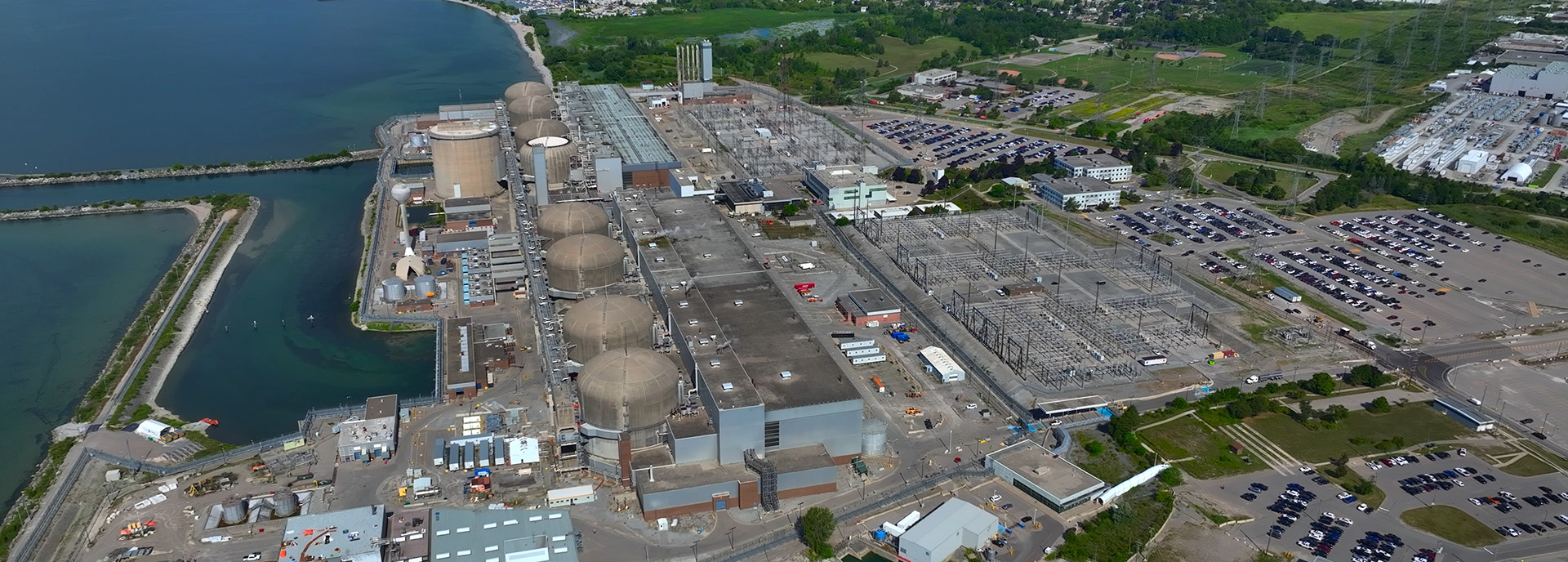 Aerial view of a large nuclear power plant facility near a body of water, with multiple domed reactor buildings, surrounding infrastructure, parking lots, and green areas in the background.