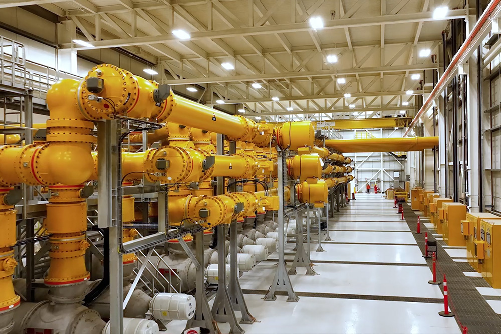 A large industrial facility with rows of yellow pipes, valves, and machinery inside a spacious, well-lit building. Orange safety cones line the walkway, and metal platforms and equipment are visible throughout.