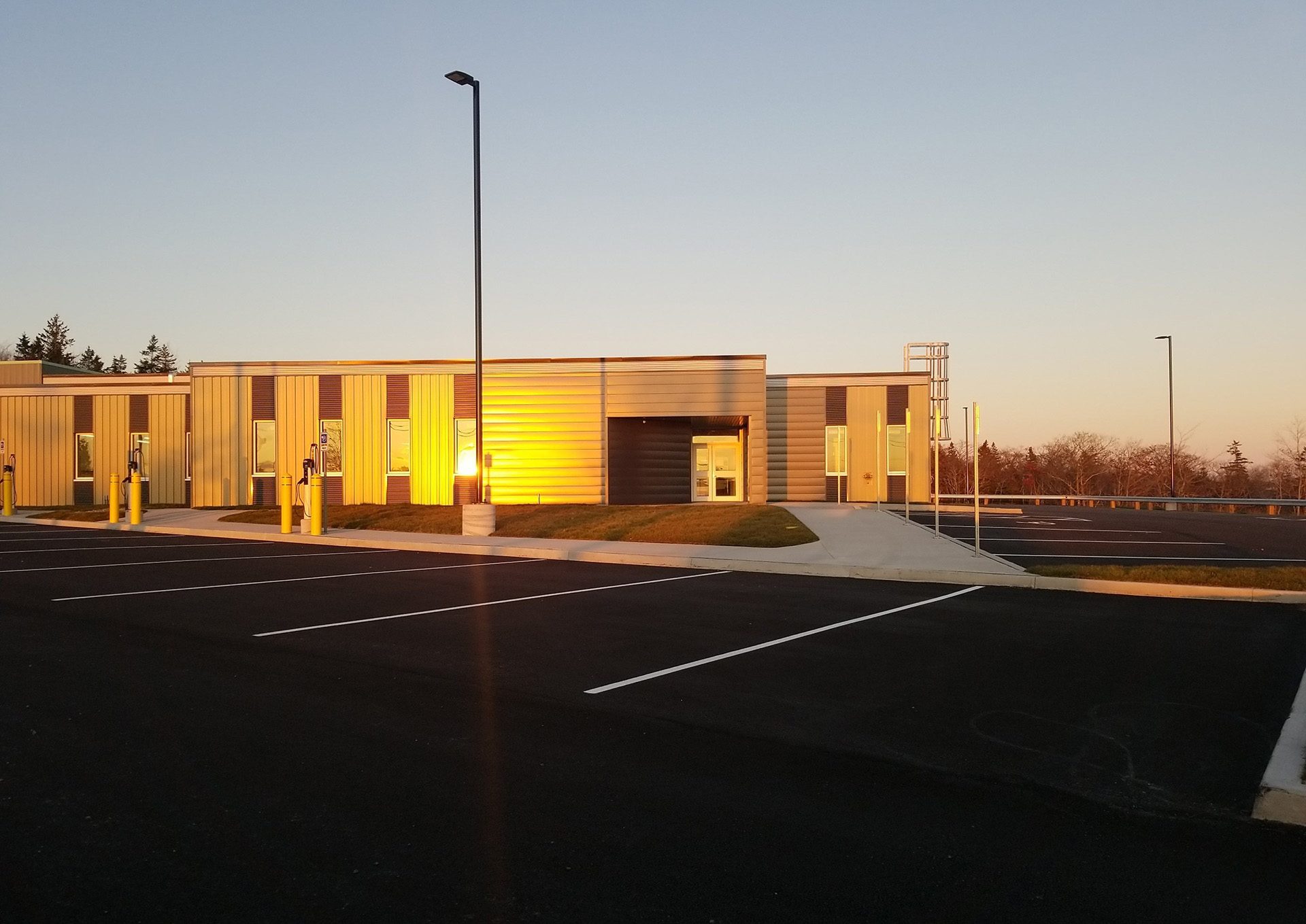 A modern, single-story building with metal siding is shown at sunset. Sunlight reflects off the windows. The empty parking lot in front has marked spaces and tall light poles. Trees are visible in the background.