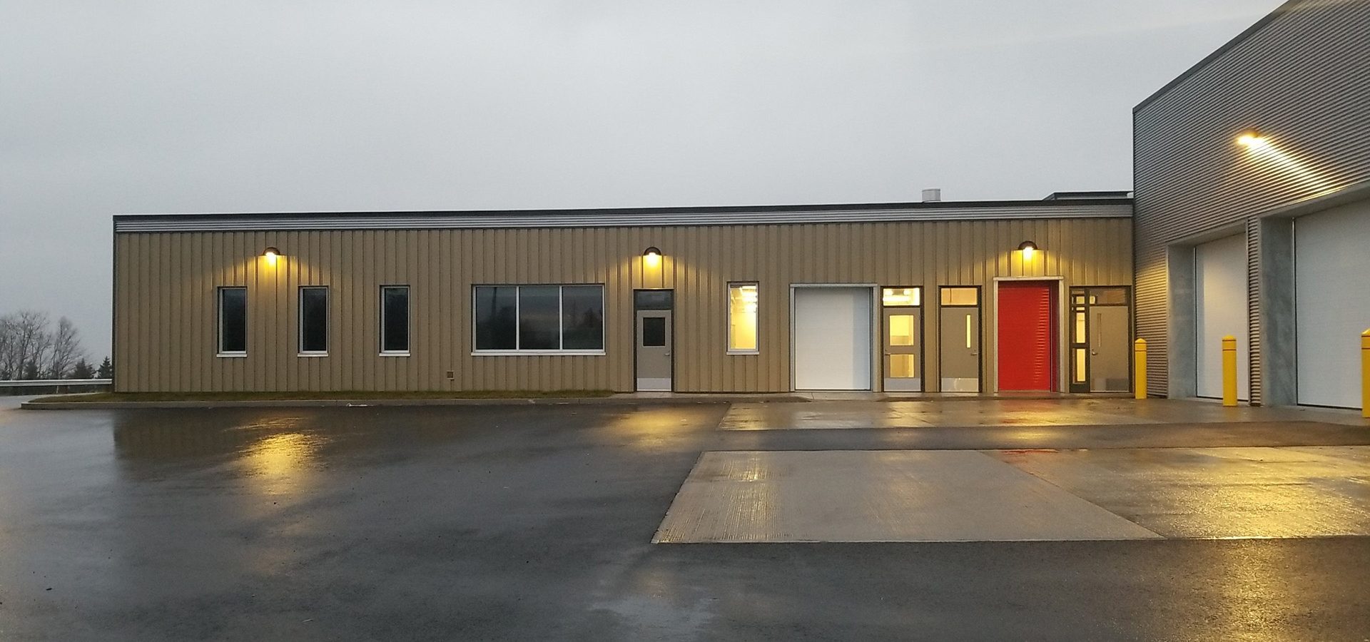 A modern industrial building with beige metal siding, several lit windows, a red door, and wet pavement reflecting the lights on a cloudy day.