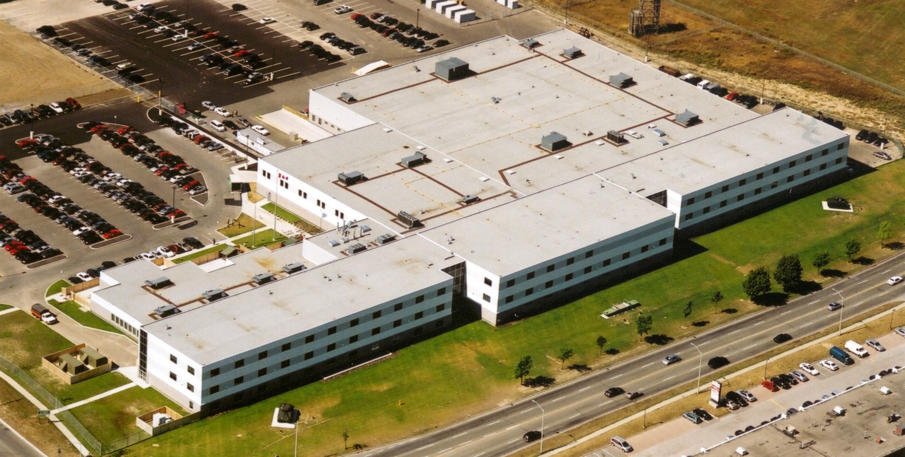 Aerial view of a large, white, rectangular building with a flat roof, surrounded by parking lots with cars and some green grassy areas, adjacent to busy roads and commercial buildings.