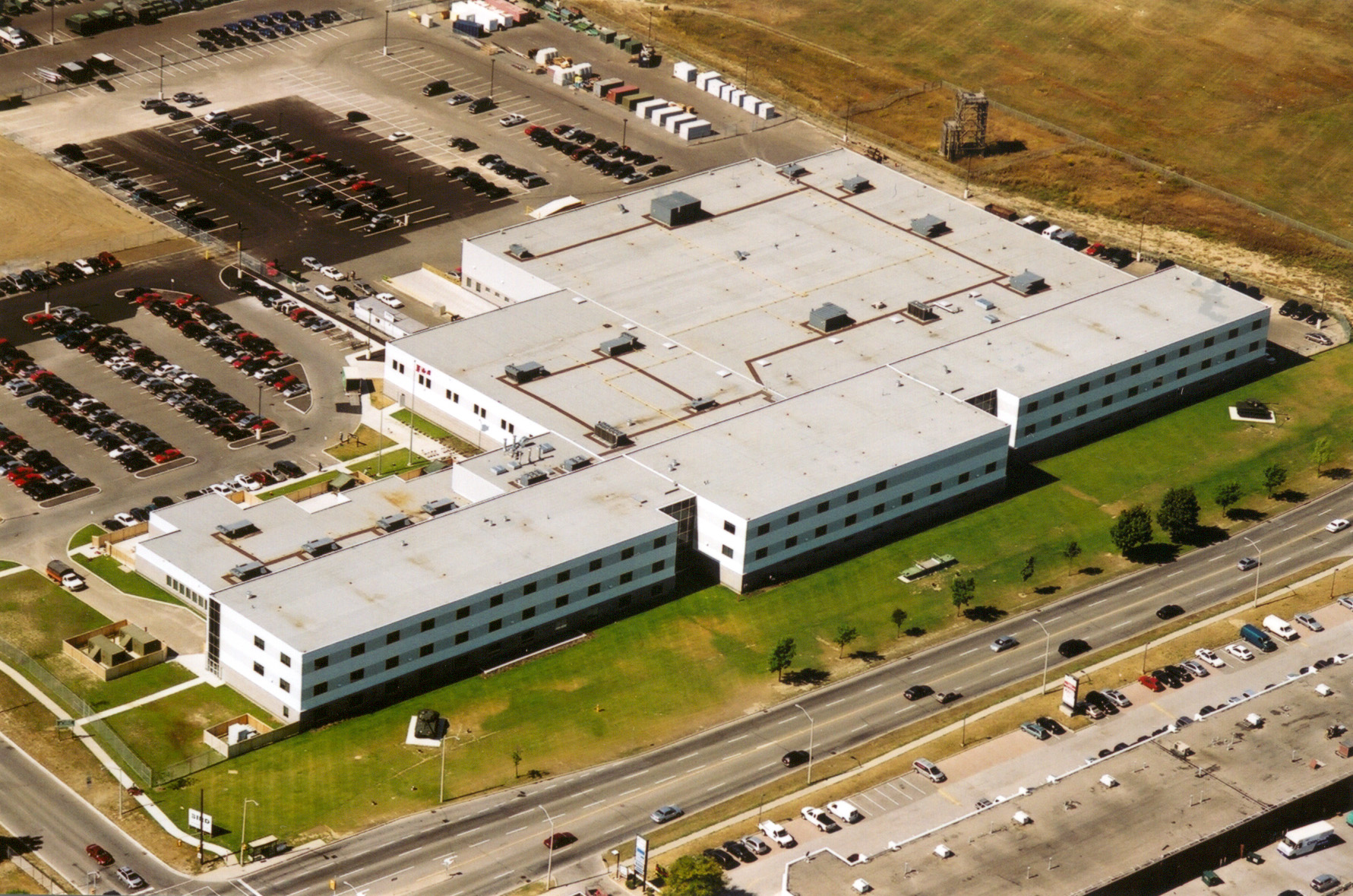 Aerial view of a large, white, rectangular building with a flat roof, surrounded by parking lots with cars and some green grassy areas, adjacent to busy roads and commercial buildings.
