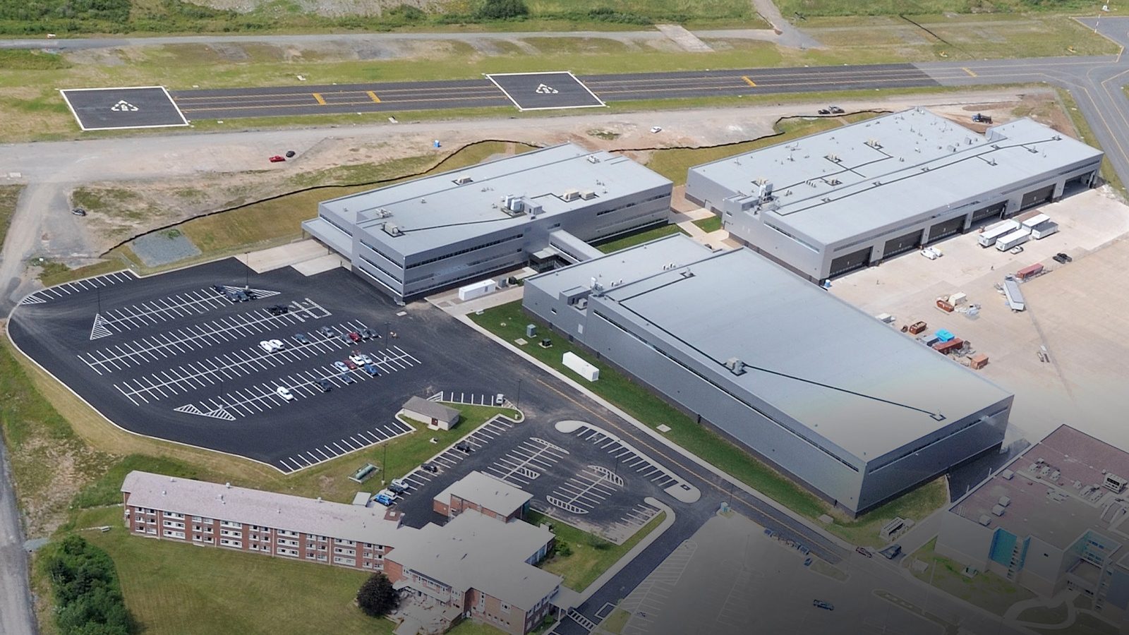 Aerial view of a large industrial complex with three main gray buildings, adjacent parking lots, and a nearby runway, surrounded by grass and additional smaller structures.