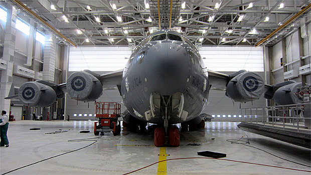 A large military cargo plane is parked inside a spacious, well-lit hangar. Workers and equipment surround the aircraft, and its engines display round insignias with a maple leaf.