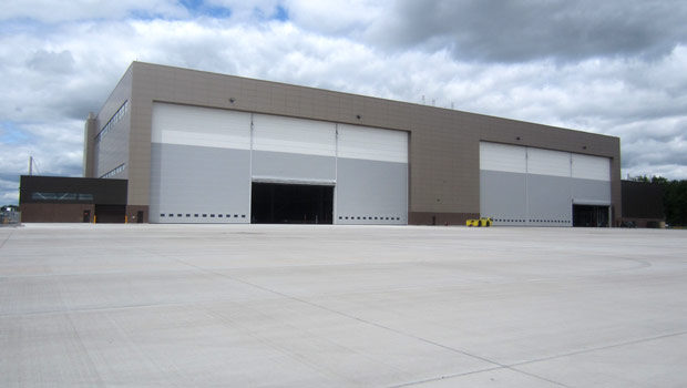 Large, modern industrial warehouse with three wide white garage doors, set on an expansive, empty concrete lot under a cloudy sky.