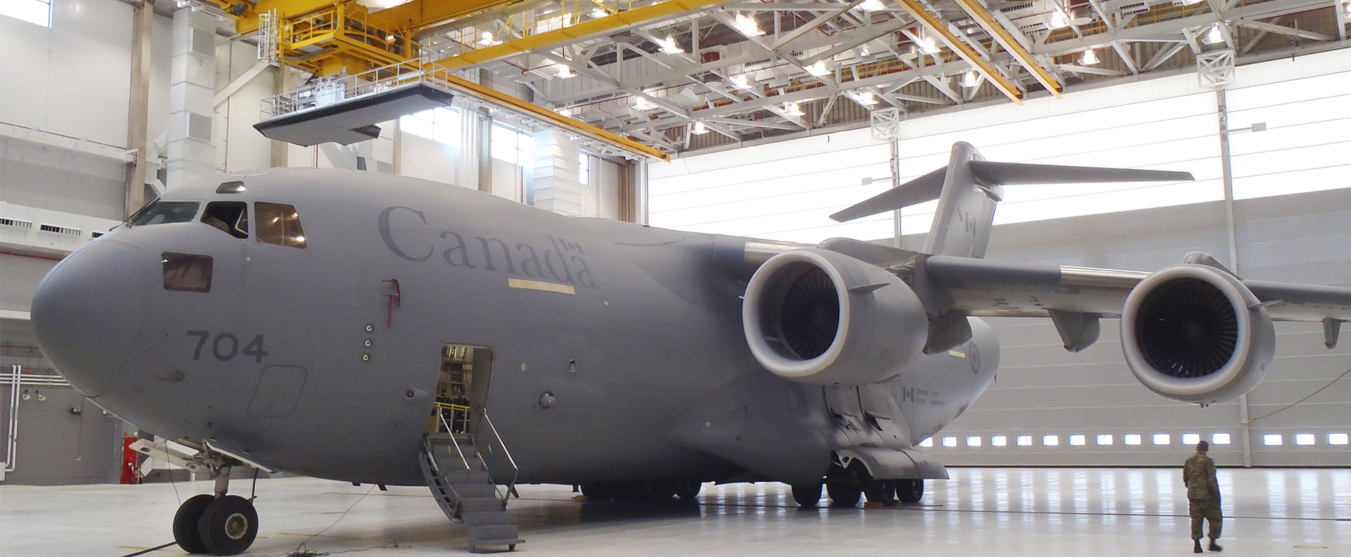 A large grey Canadian military cargo plane, marked Canada and number 704, is parked inside a spacious hangar with its stairs down. A person in uniform stands nearby for scale.