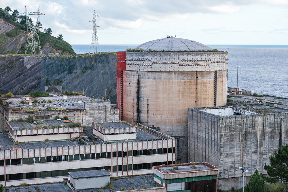 An abandoned nuclear power plant with a large concrete containment dome, overgrown vegetation on rooftops, and rusted structures, situated near the sea with hills and power lines in the background.