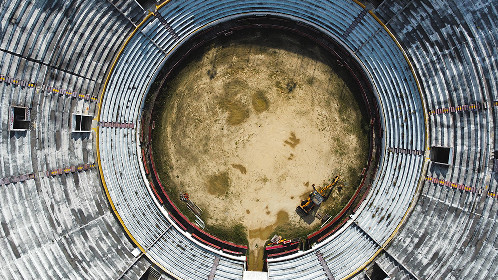 Aerial view of an empty circular arena with tiered seating and a dirt-covered center, where a small yellow construction vehicle is parked near the edge of the arena floor.