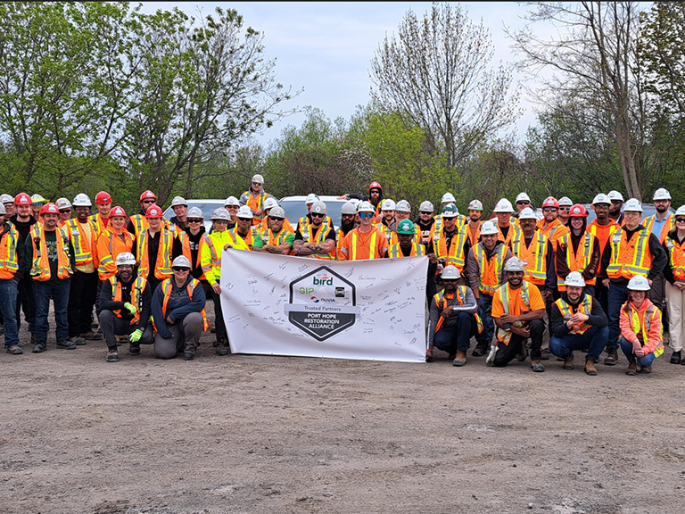 A large group of construction workers wearing hard hats and safety vests pose outdoors, holding a signed banner with logos and text. Trees and overcast sky are visible in the background.