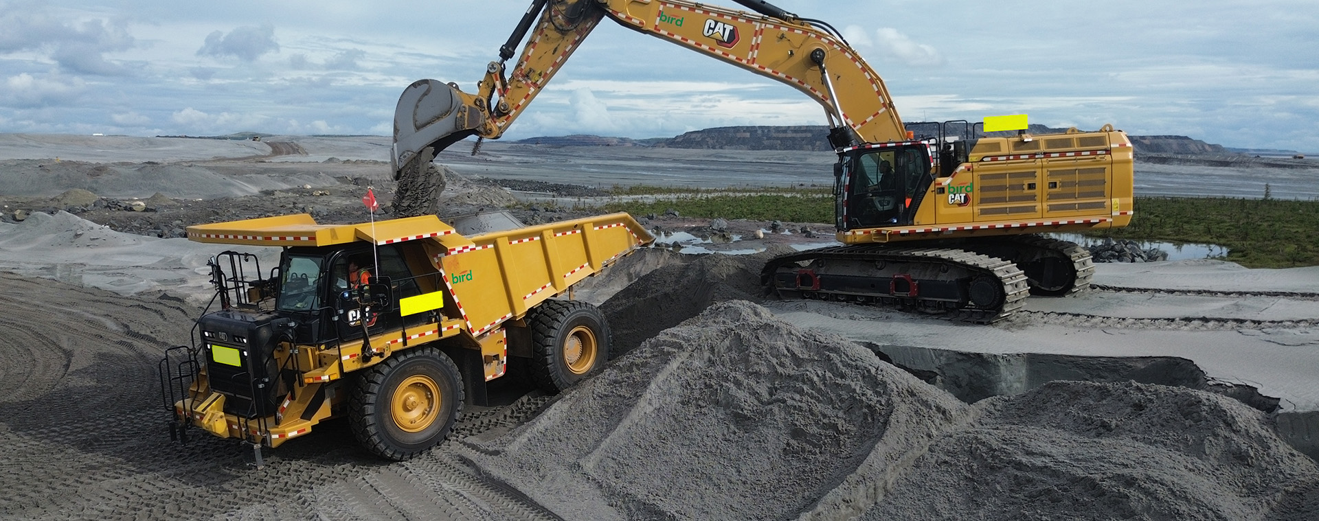 A large excavator loads dirt into a dump truck at a mining or construction site, with piles of earth and a cloudy sky in the background.
