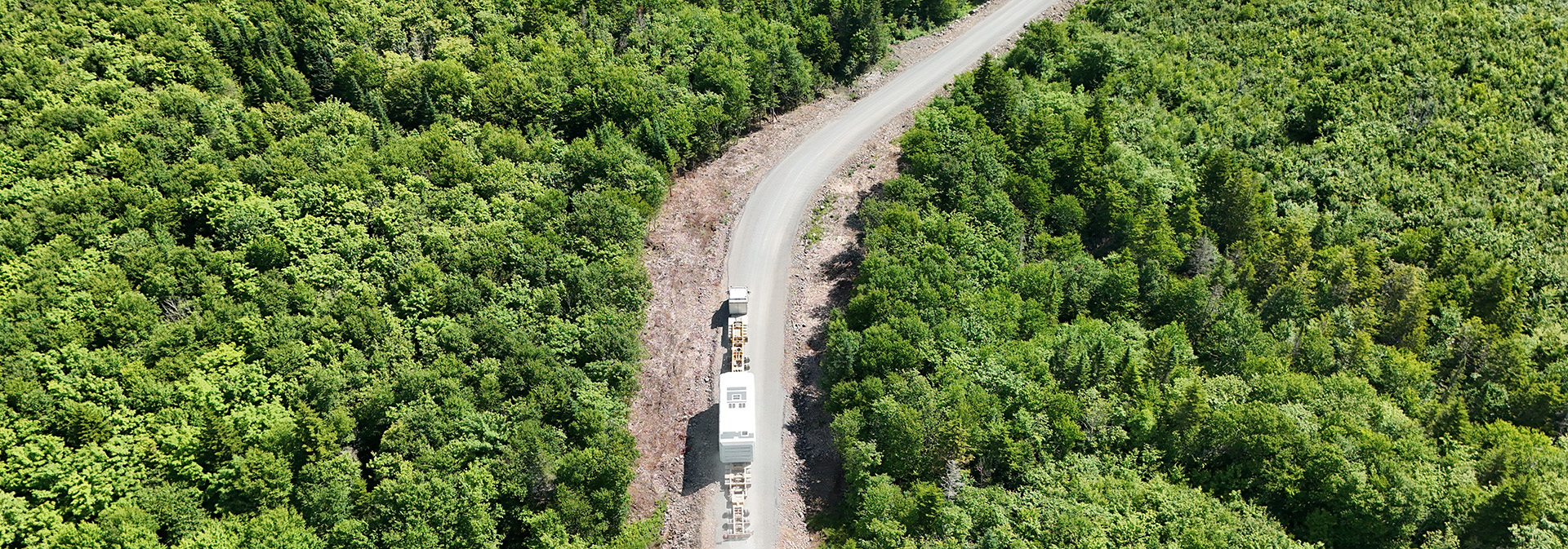 Aerial view of a white truck driving along a winding road surrounded by dense green forest.