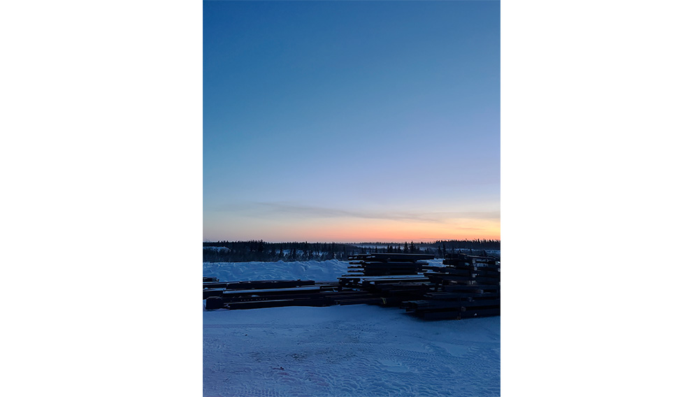 Snow-covered ground with stacks of wooden beams in the foreground. A forested tree line is visible in the distance under a clear blue sky at sunset, with orange and purple hues near the horizon.