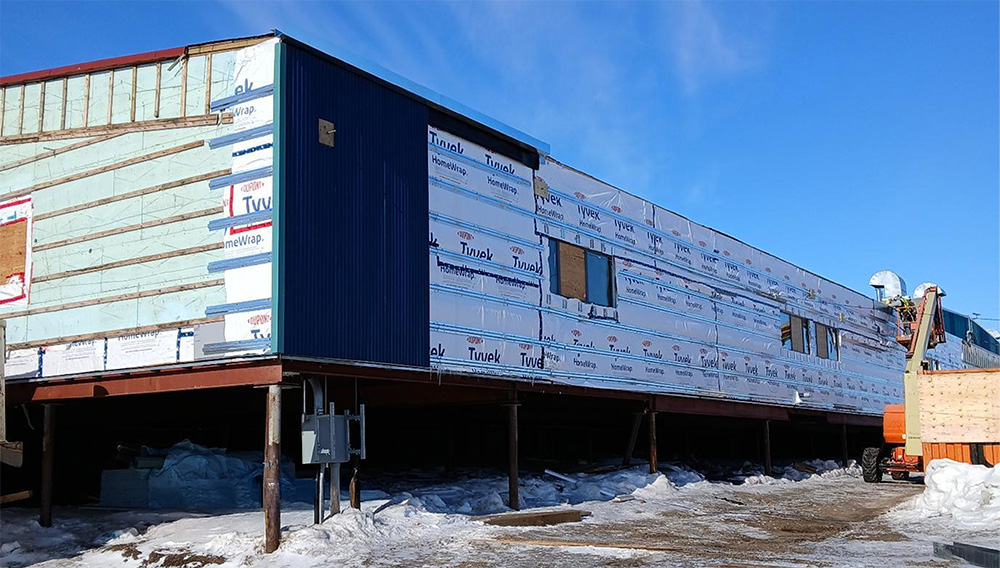 A long, rectangular building under construction stands on stilts above snowy ground. The exterior is partially covered with insulation panels and building wrap, set against a clear blue sky.