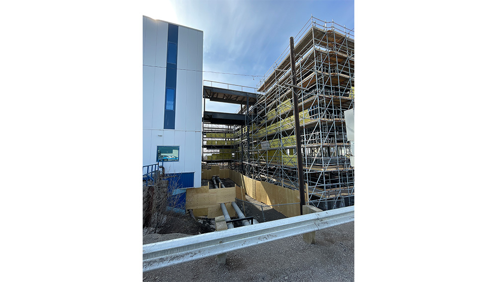 Construction site with metal scaffolding next to a large building. Snow covers the ground, and wooden barriers line the walkway. A skybridge connects two structures above the scaffolding.