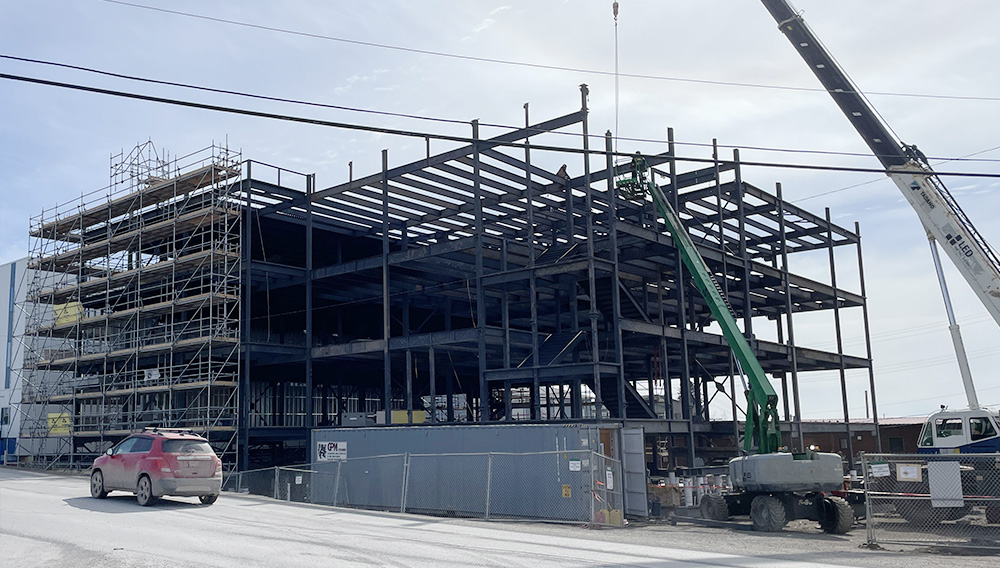 A construction site showing the steel framework of a multi-story building in progress, with construction materials and equipment on the ground and a crane visible on the right.