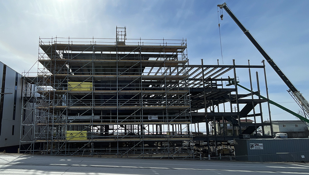 A steel framework of a multi-story building under construction, surrounded by snow and construction equipment, with a blue sky in the background.