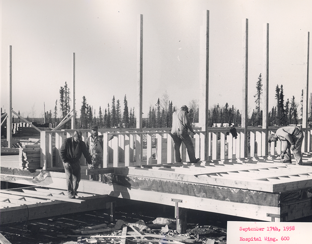 Black and white photo of four workers constructing a wooden building frame on a raised platform, surrounded by trees. A label reads: September 17th, 1958, Hospital Wing, 600.