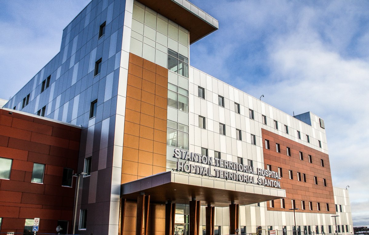 A modern, multi-story hospital building with silver, white, and brown panels. The sign reads Stanton Territorial Hospital above the main entrance. The sky is blue with light clouds.