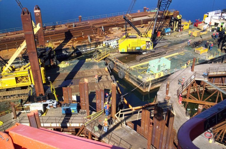 A construction site on the water features cranes, metal beams, machinery, and workers in safety gear. The area is busy with equipment and structural materials, and calm blue water is visible in the background.