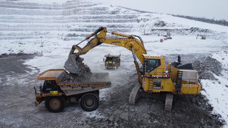 A large yellow excavator loads rocks into a dump truck at a snowy open-pit mining site with terraced slopes and overcast skies in the background.
