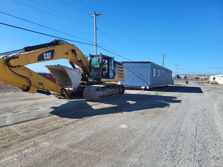 A yellow CAT excavator is parked on a gravel road with its bucket raised, next to a large blue portable building. Power lines and clear blue sky are visible in the background.