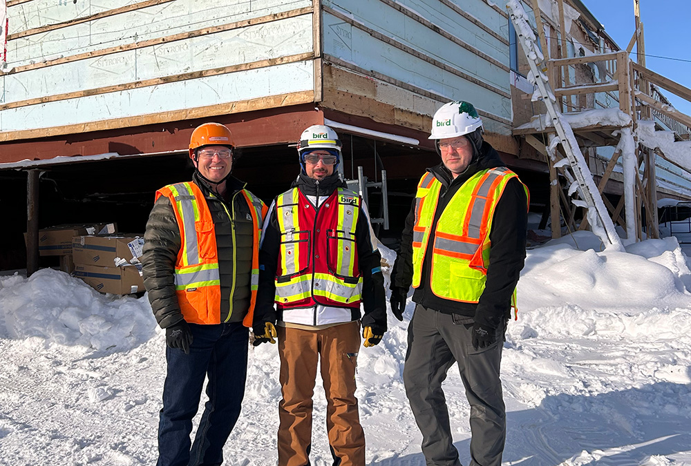 Three people wearing hard hats, winter coats, and high-visibility vests stand on snowy ground in front of a partially constructed building covered in insulation panels.