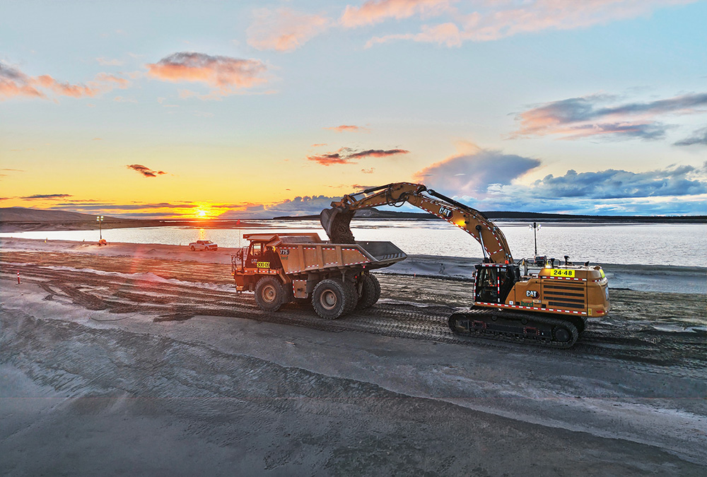 An excavator loads soil into a large dump truck on a construction site near water at sunset, with a dramatic sky and distant lights on the horizon.