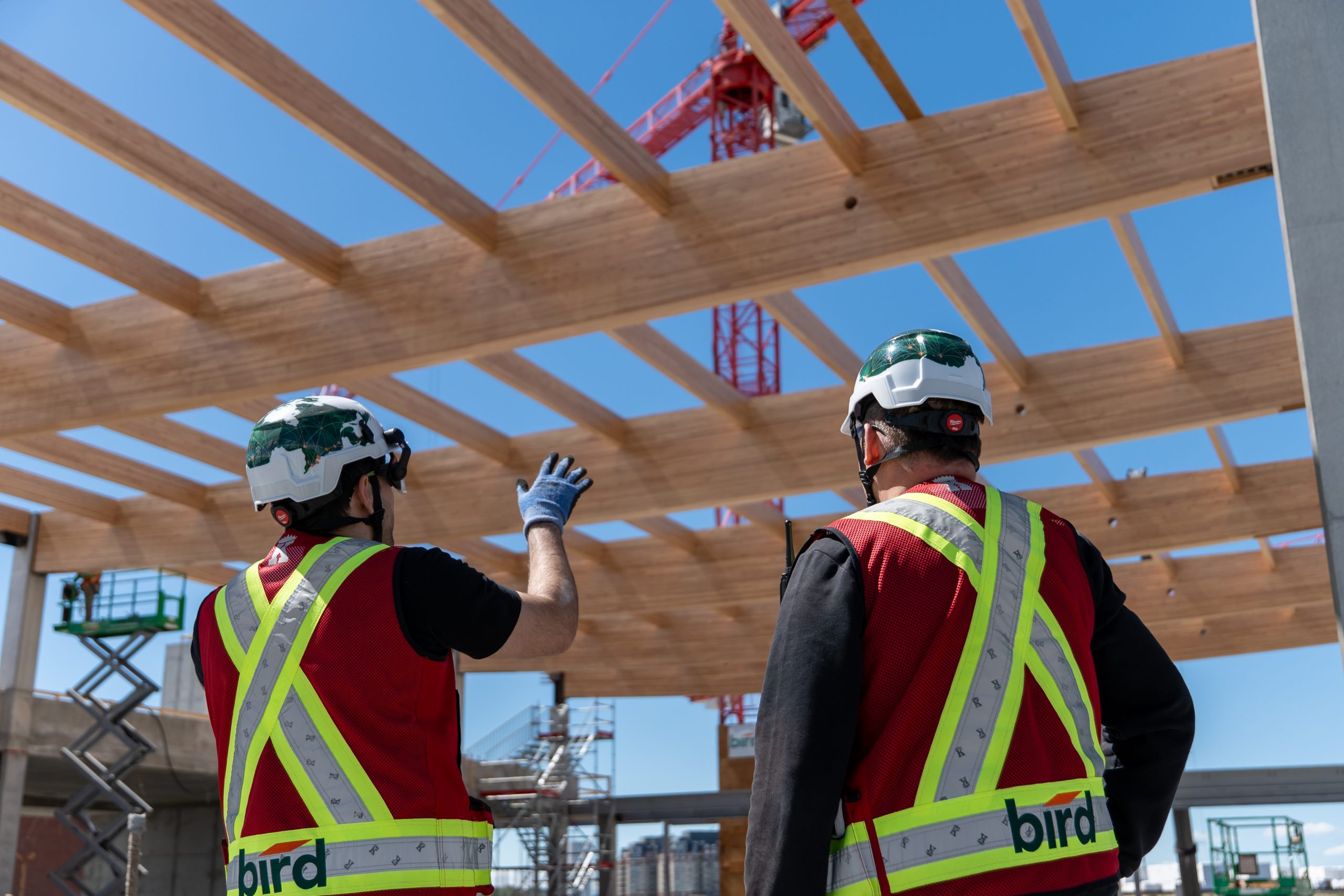 Two construction workers in safety vests and helmets discuss a wooden structure being built, with a crane and lift in the background under a clear blue sky.